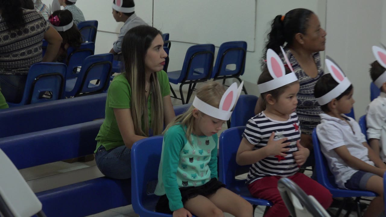 In Cabo San Lucas, three children in the front row sit quietly in blue chairs as a woman in a green shirt looks toward the room.