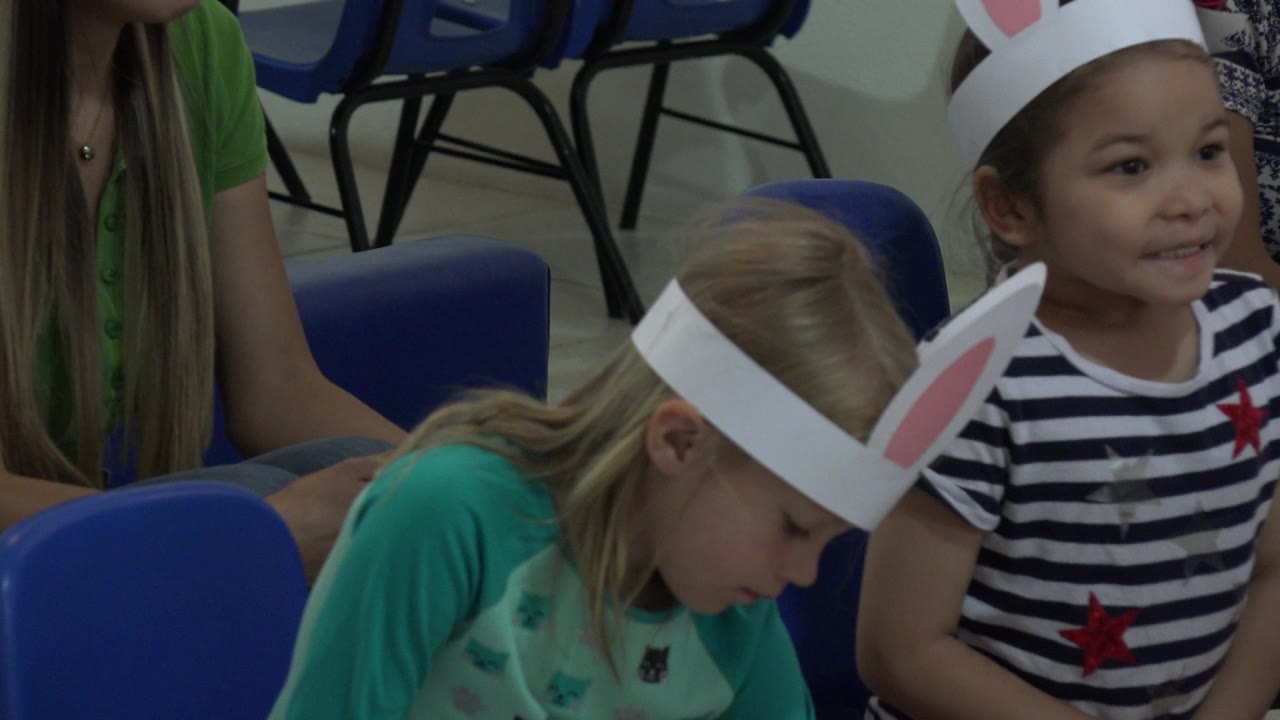 Two young girls in paper bunny ears sit among blue chairs.