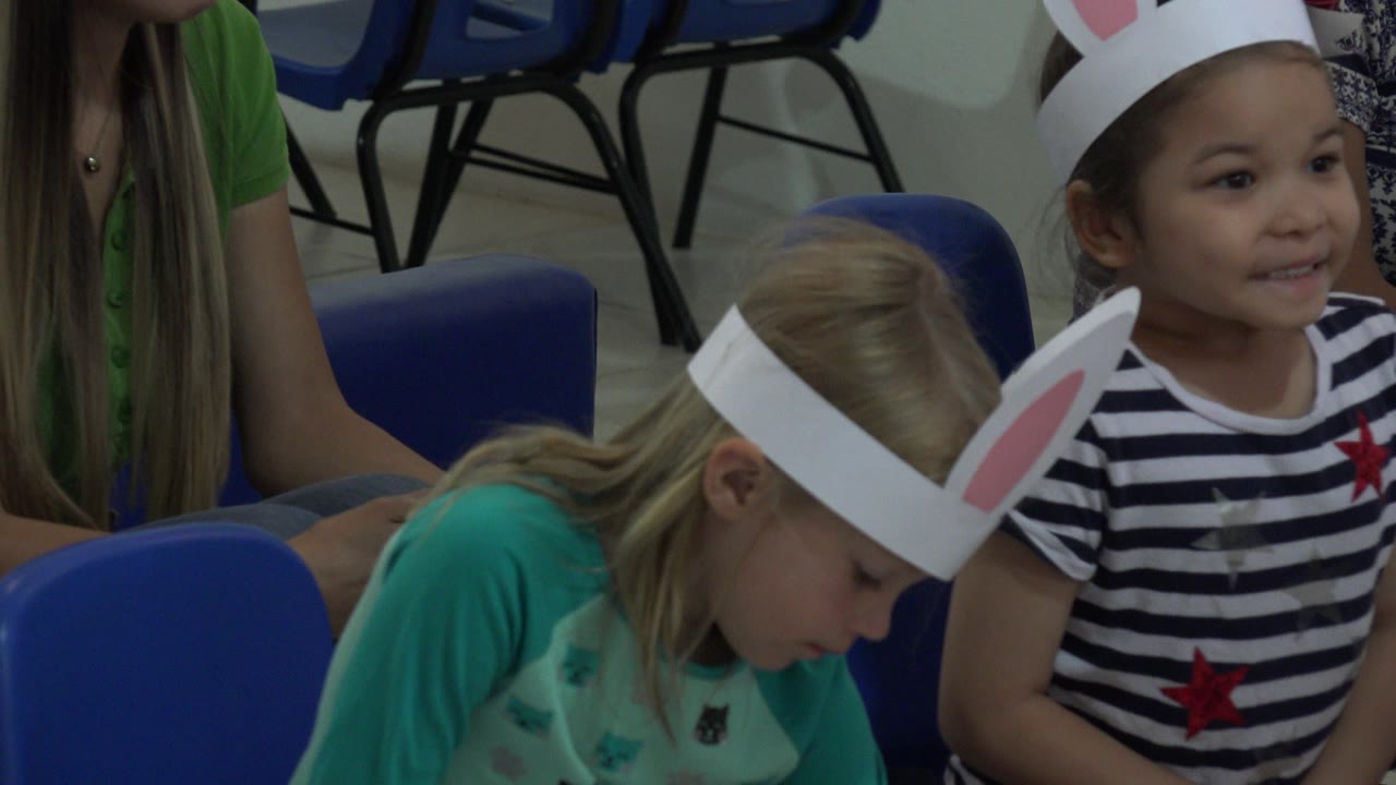 A smiling child in a striped shirt and white bunny ears sits beside a blonde child looking down in blue chairs in Cabo San Lucas.