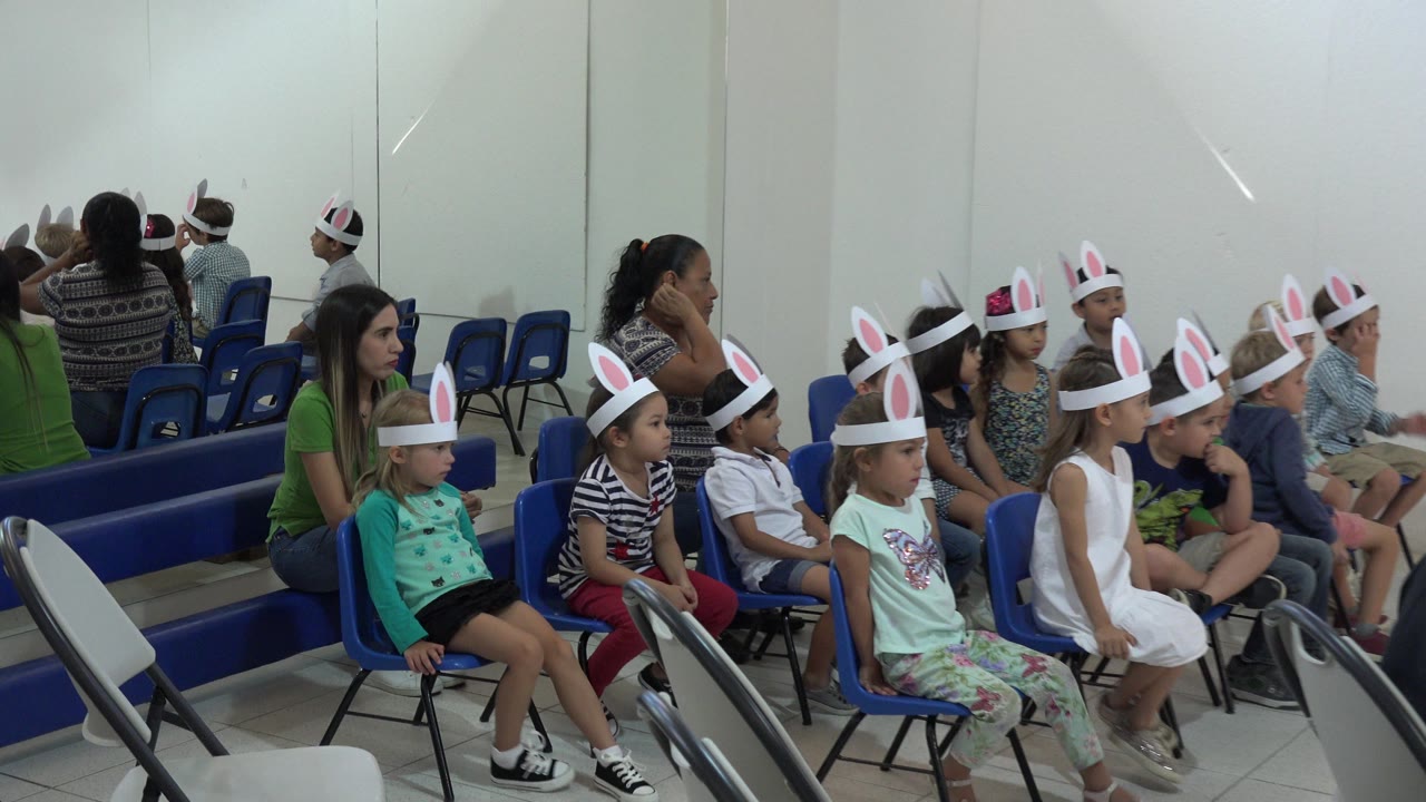 Children in paper bunny-ear headbands sit in a row of blue chairs during an indoor Easter gathering in Cabo San Lucas, Mexico, with adults seated nearby.