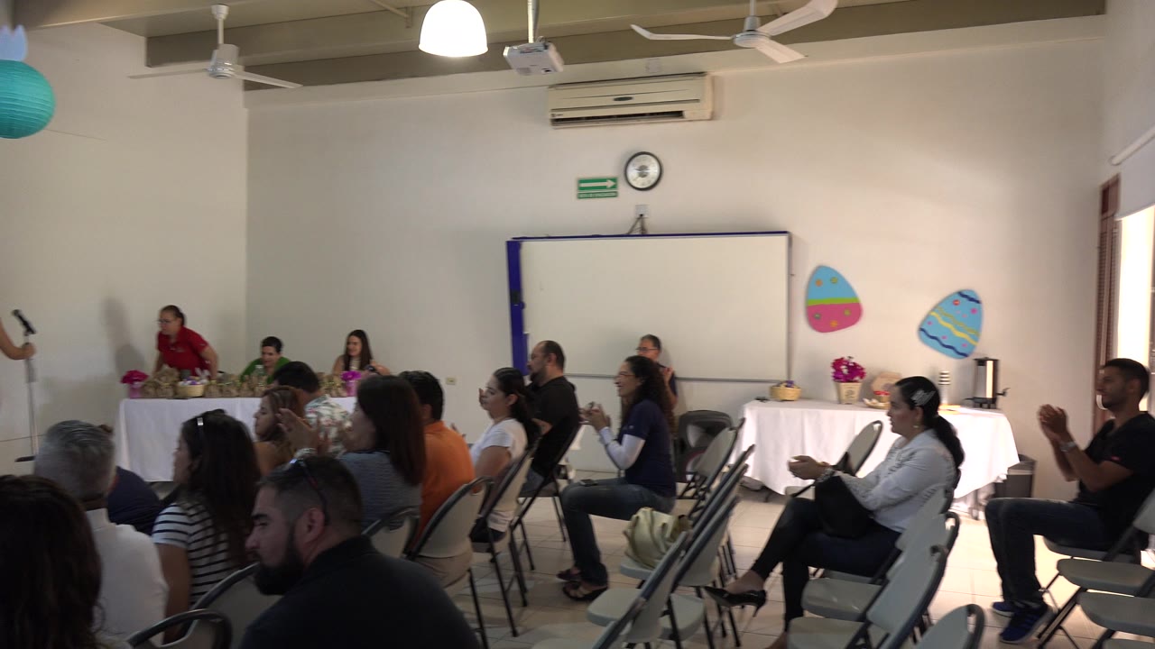 Seated adults in Cabo San Lucas, Mexico clap toward an off-camera presentation, with rows of gray folding chairs, a whiteboard, and ceiling fans visible.