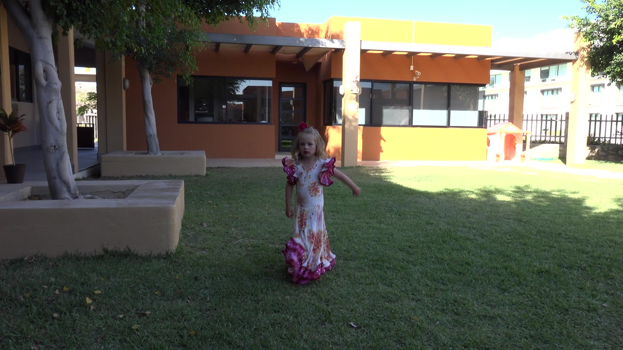A young girl in a floral dress with pink ruffled sleeves walks across grass in a shaded courtyard in Cabo San Lucas, Mexico, with a tan and orange building behind her.