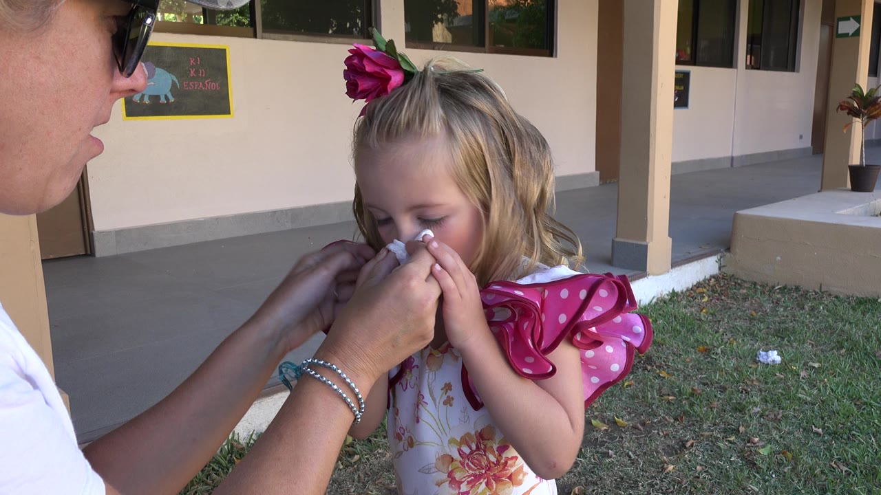 In Cabo San Lucas, Mexico, a woman at the left edge uses a tissue on a young blonde girl's nose while the child stands in a pink ruffled dress outside a beige building.
