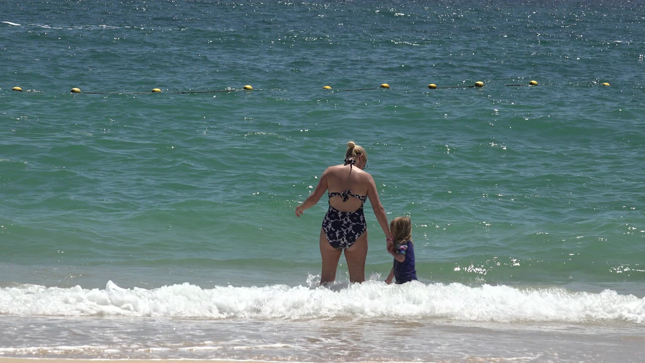 Mother and young daughter holding hands in shallow surf.