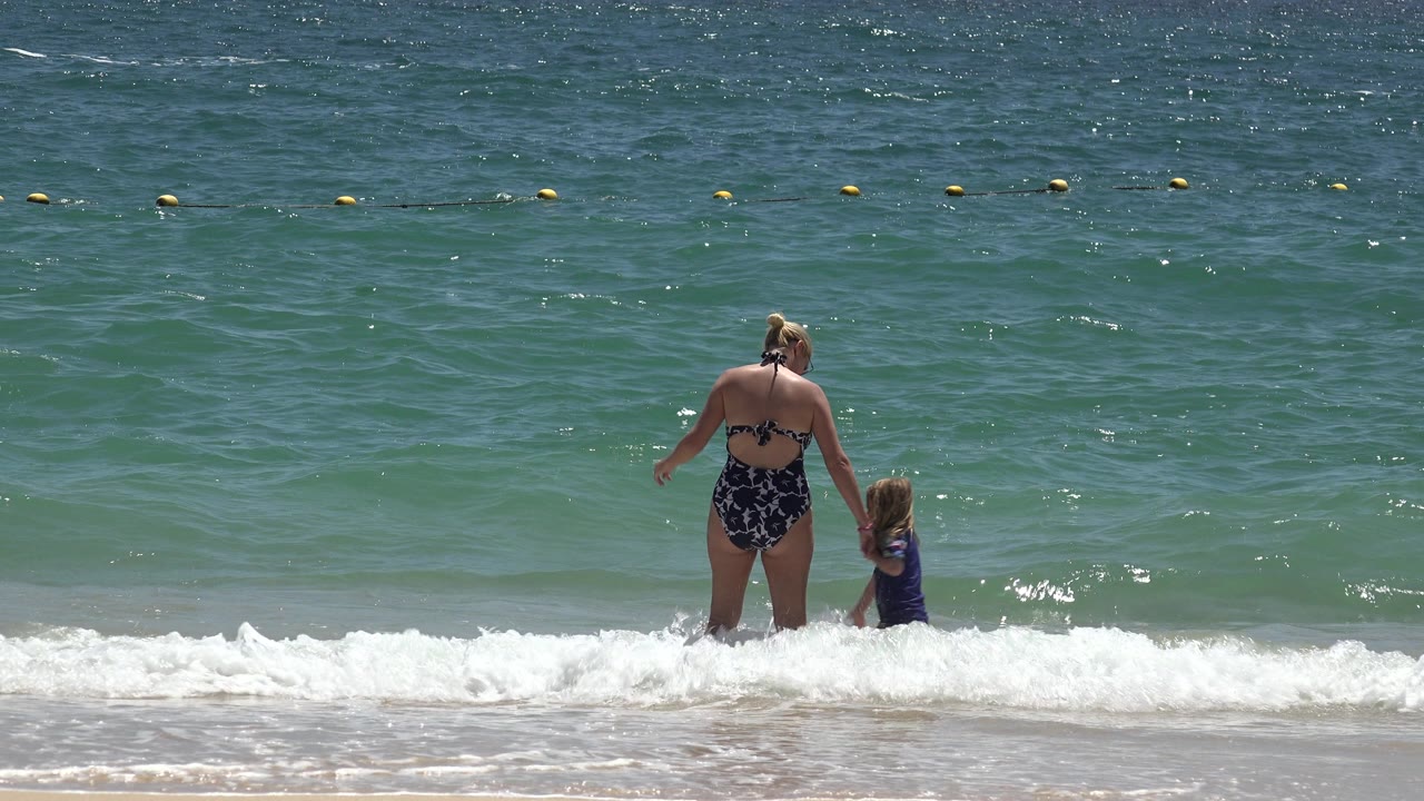 The mother and daughter remain near the waterline in Cabo San Lucas while a low white wave spreads across the foreground sand.
