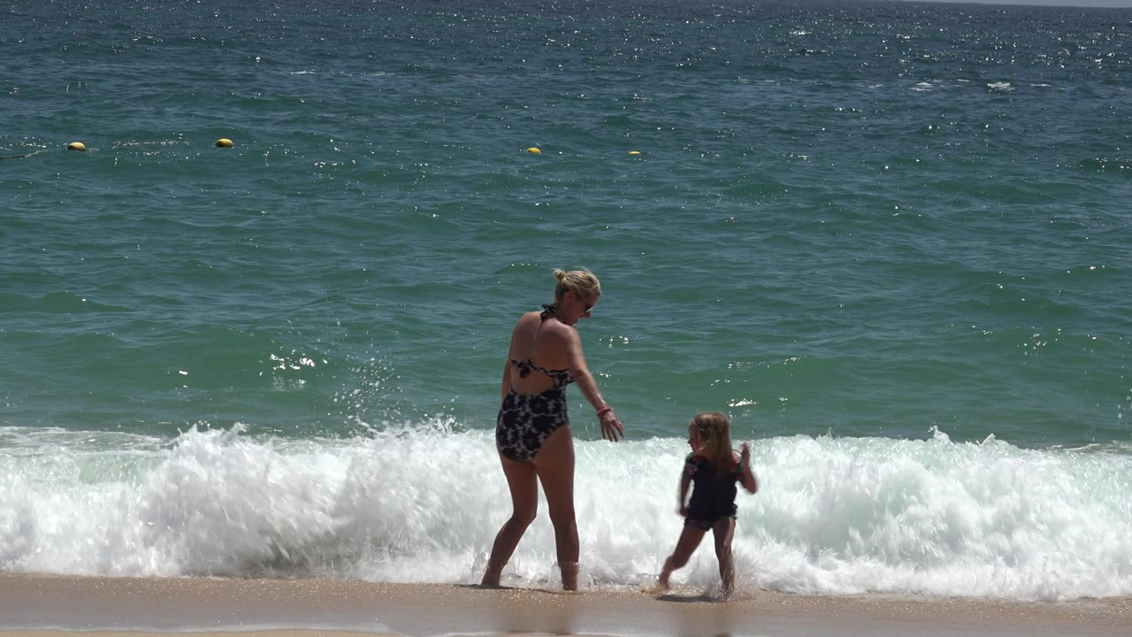 Adult woman in a dark patterned swimsuit and young child in a dark swim shirt stand near breaking waves on a Cabo San Lucas beach.