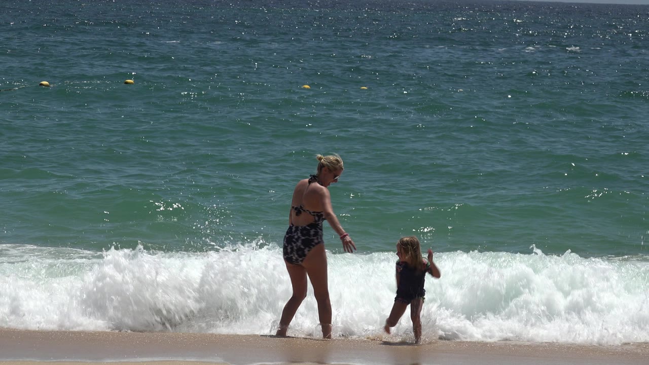 Adult woman faces a young child as a fresh white wave collapses behind them on the Cabo San Lucas shoreline.