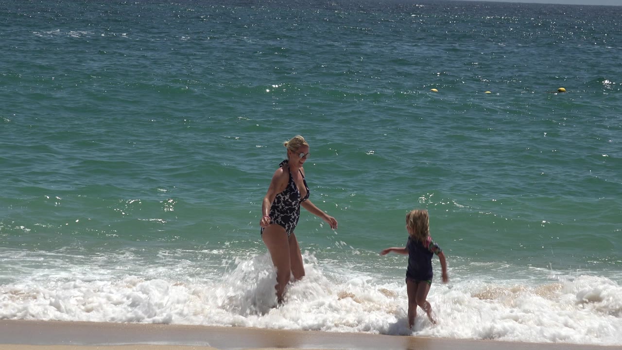Woman and child walk through churning foam at the beach.