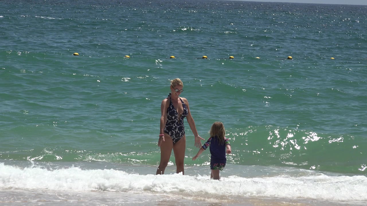 Woman and young girl stand in shallow surf on a sunny beach.