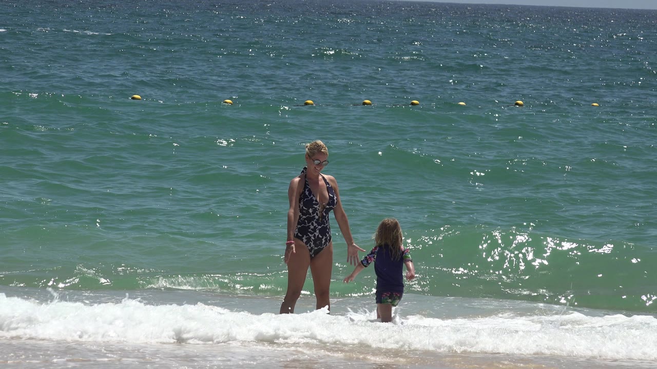 The woman and young girl hold hands in the shallow water at Cabo San Lucas as sunlight reflects on the small wave behind them.
