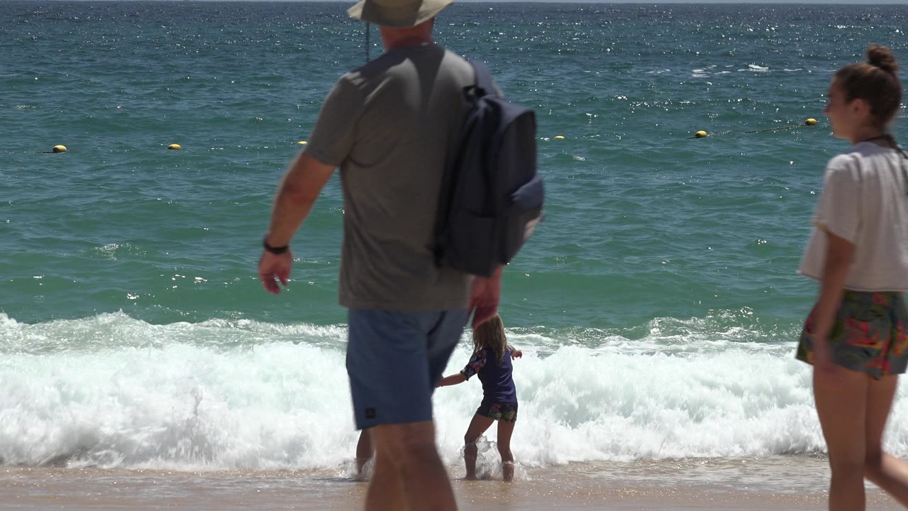 At a Cabo San Lucas beach, a man with a backpack and a young woman in shorts pass in the foreground while a woman holds a child in the foamy surf behind them.