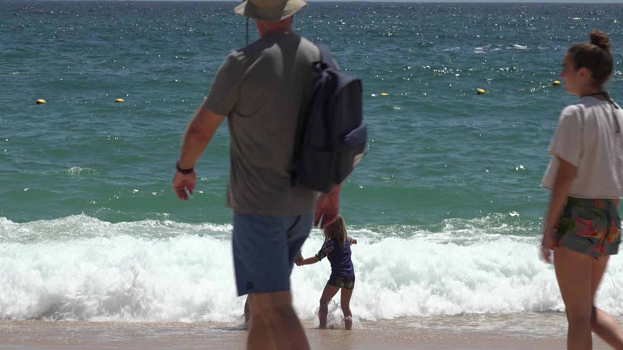 The Cabo San Lucas shoreline remains framed from ground level as foreground beachgoers walk across the shot and the woman and child stay near breaking whitewater.