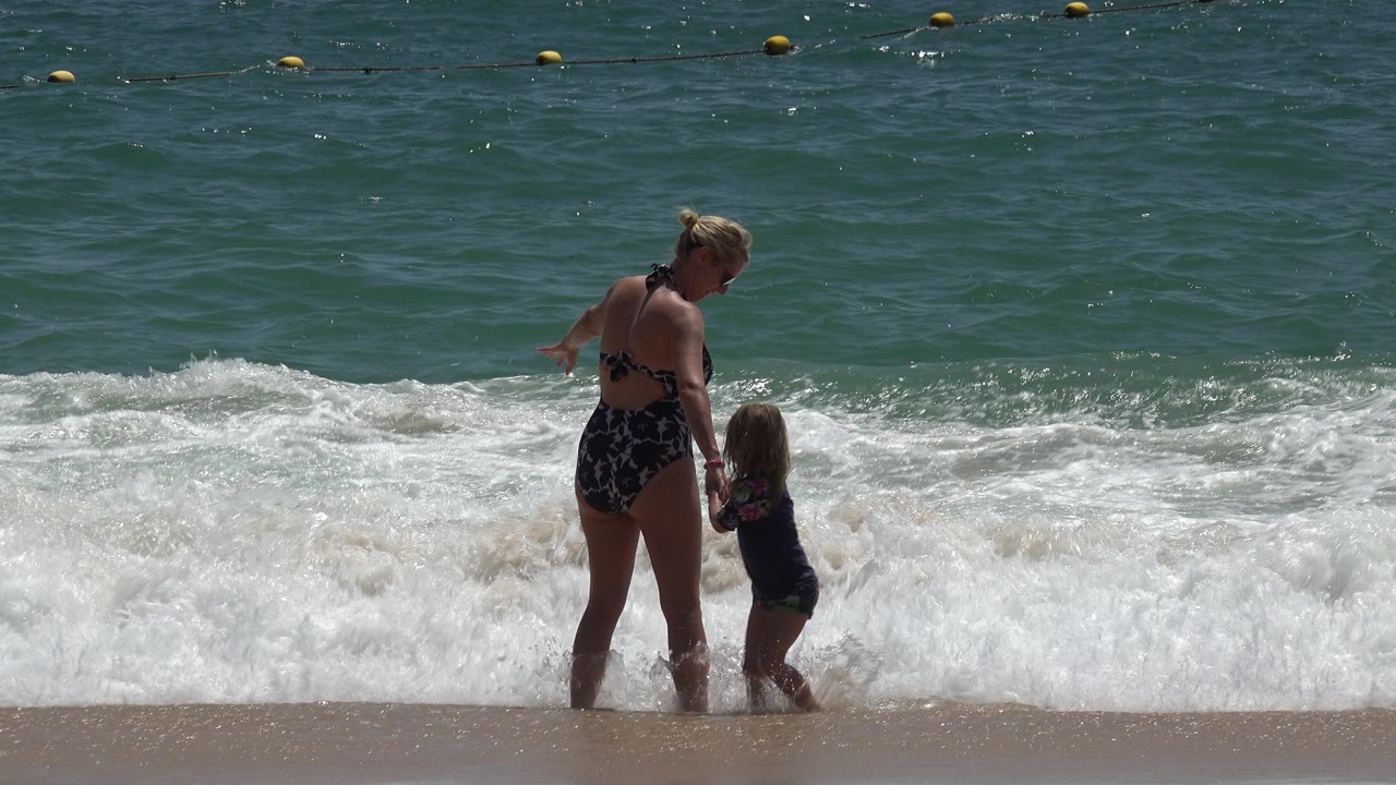 Adult woman holding a child's hand at the edge of breaking waves.