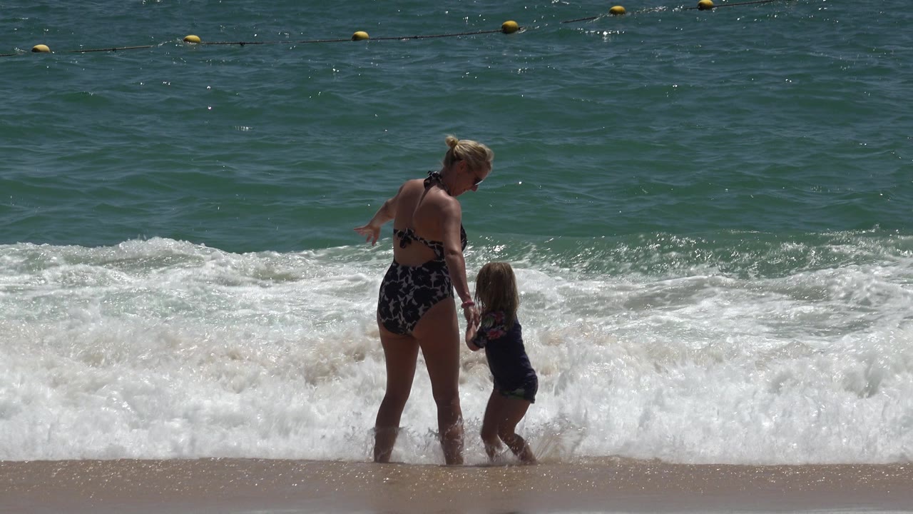 In Cabo San Lucas, Mexico, the woman and child pause in knee-high foam as a wave rolls in behind them under bright sun.