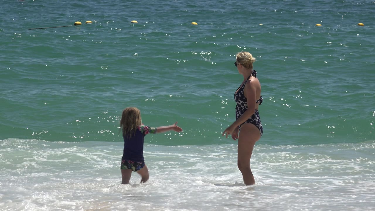 A child faces an adult woman in shallow surf at Cabo San Lucas, Mexico, with white foam at their legs and green-blue ocean filling the background.