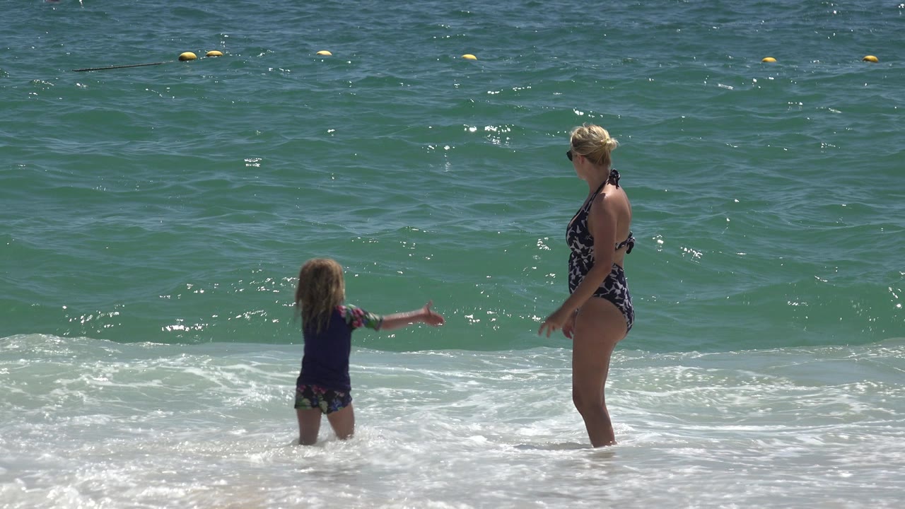 The child extends an arm toward the adult woman in the Cabo San Lucas surf as sunlight reflects on the water and a buoy line sits far behind them.