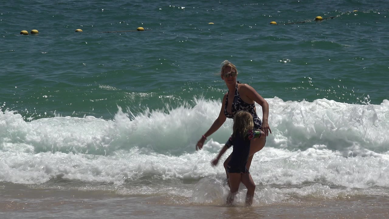 A mother in a dark patterned swimsuit stands beside a young child at the Cabo San Lucas shoreline as a white wave breaks behind them.
