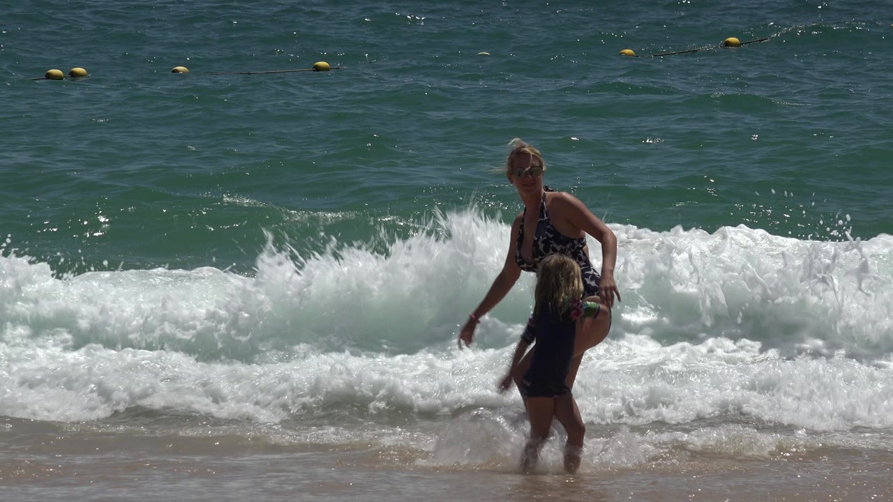 The child turns near the woman at the Cabo San Lucas waterline while foamy surf rolls across the sand behind their legs.