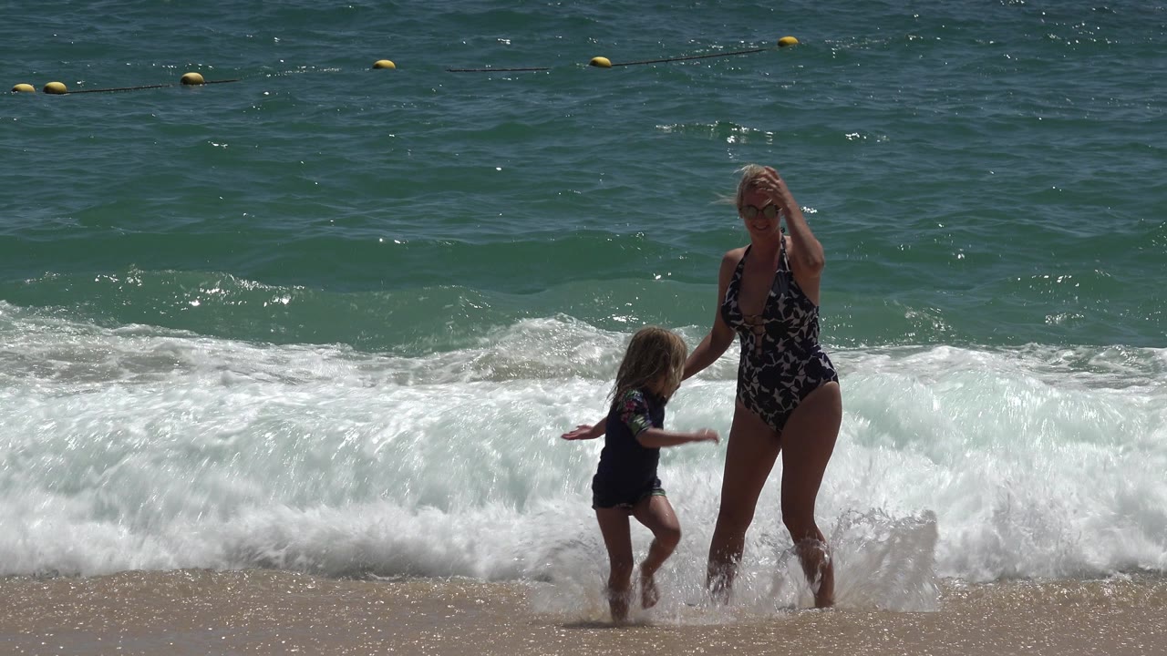 Child walks beside woman as a wave washes onto the beach.