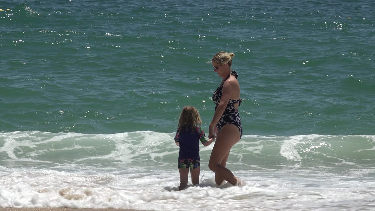The woman and child remain hand in hand in Cabo San Lucas, Mexico, facing the shore break as sunlight reflects across the textured ocean surface.