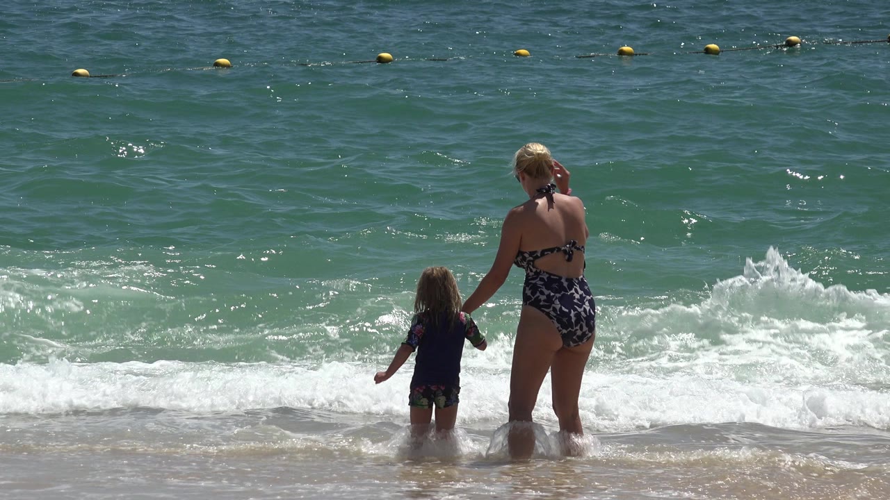 Woman and small child stand at the edge of ocean surf.