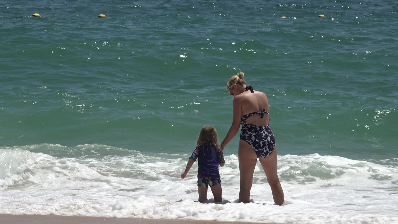 In Cabo San Lucas, Mexico, a woman in a patterned swimsuit holds a child's hand as both stand ankle-deep at the foamy edge of the ocean.
