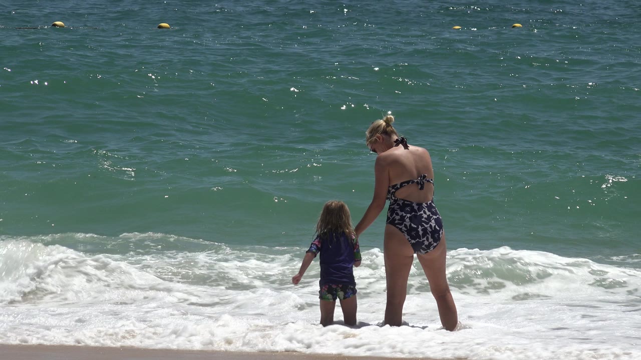 In Cabo San Lucas, Mexico, the woman leans her attention toward the child while a low white wave approaches their legs from the ocean.