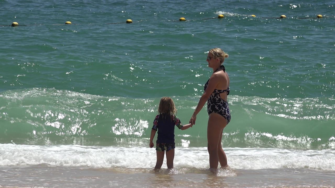Woman and child holding hands at the shoreline as a small wave approaches.