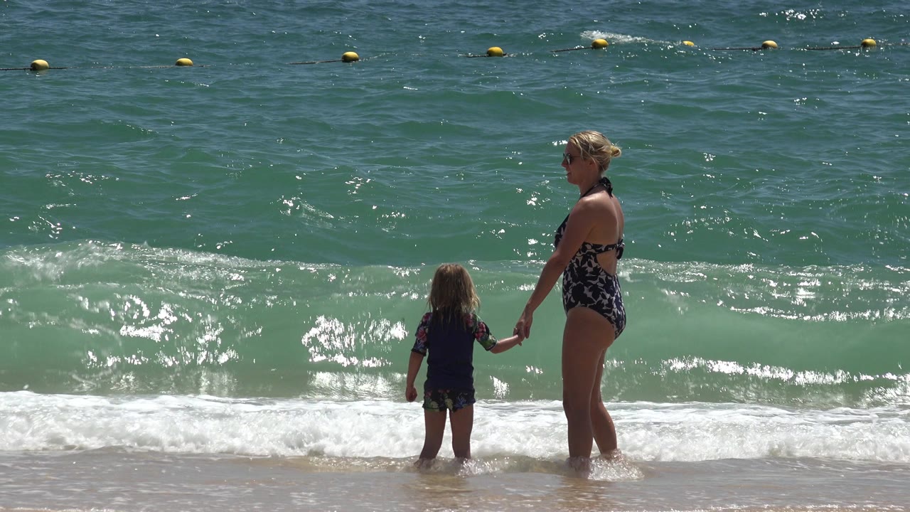 In Cabo San Lucas, Mexico, the woman and child remain hand in hand near the waterline as sunlight flashes across the darker ocean surface.