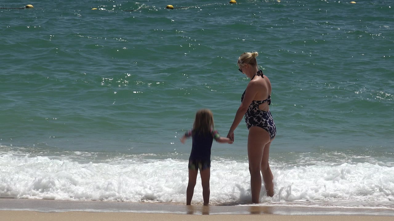 In Cabo San Lucas, Mexico, a woman in a patterned swimsuit holds a small child's hand as they face the incoming surf near the sandy shore.