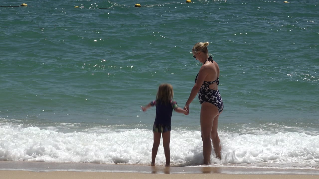 In Cabo San Lucas, Mexico, the woman and child remain hand in hand while white foam spreads across the beach in front of them.