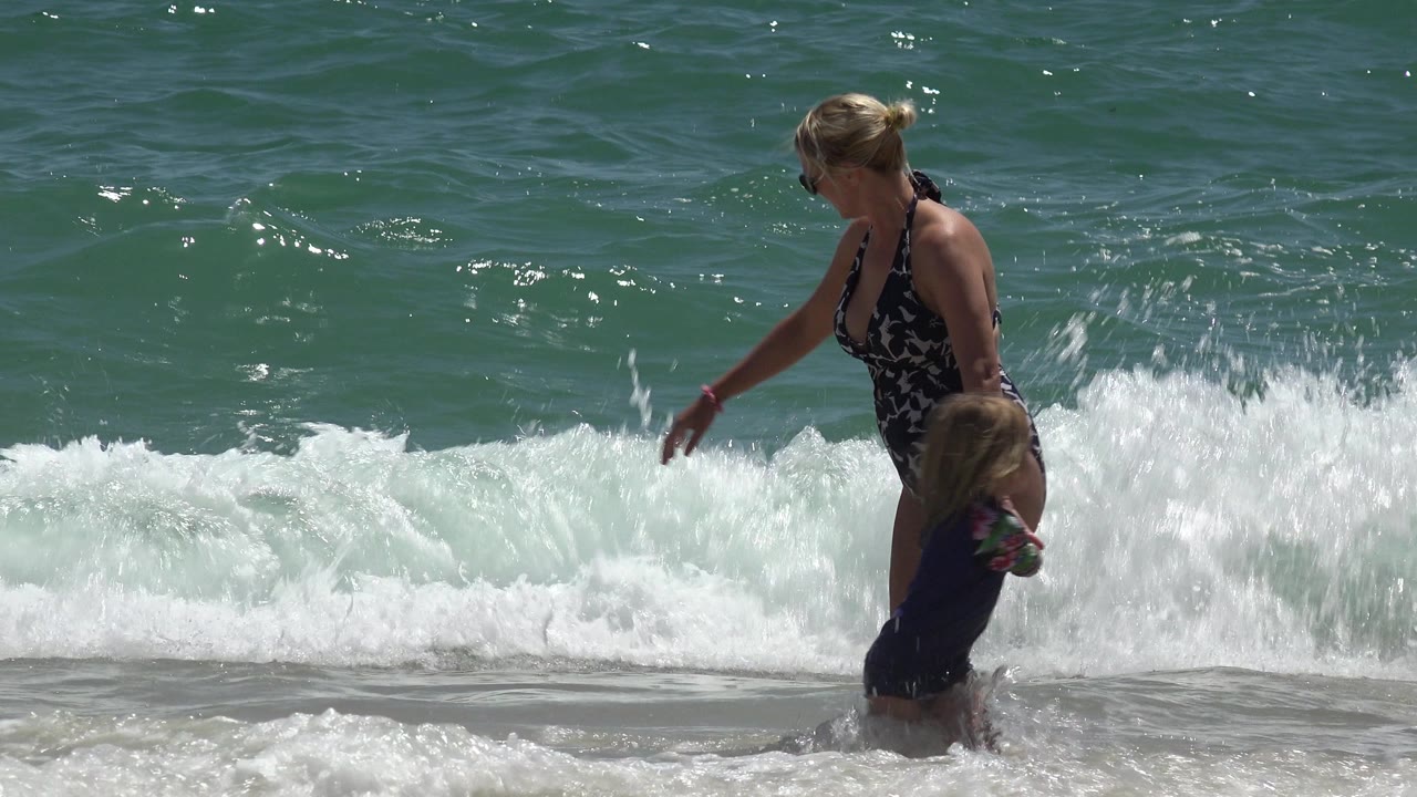 In Cabo San Lucas, Mexico, a woman reaches toward a young child in shallow beach water while a white wave breaks behind them.