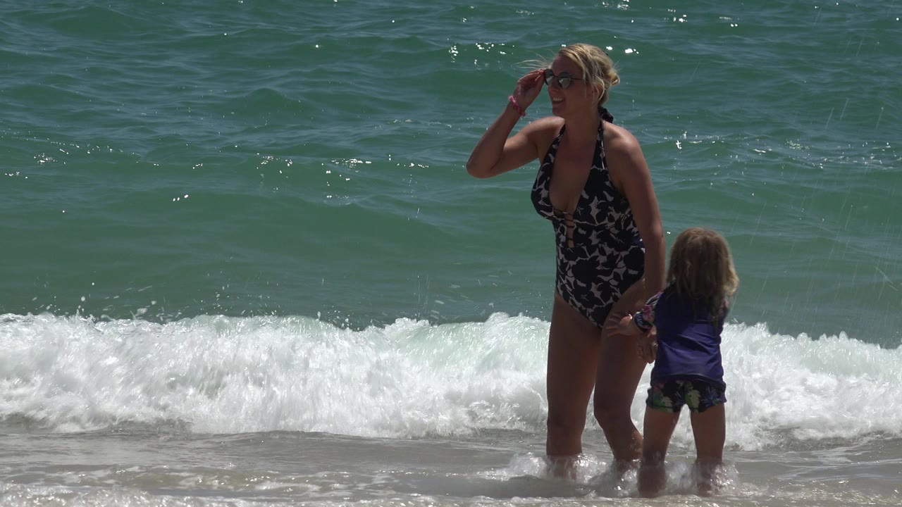 Woman adjusts sunglasses while holding a child's hand at the beach.