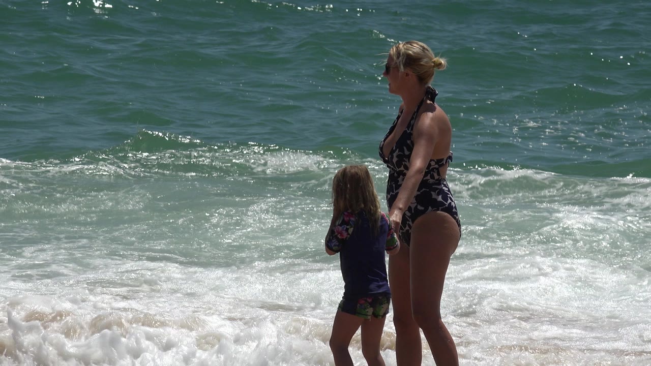 The woman and child remain near the surf in Cabo San Lucas, with the child facing the water and a white wave spreading across the lower frame.
