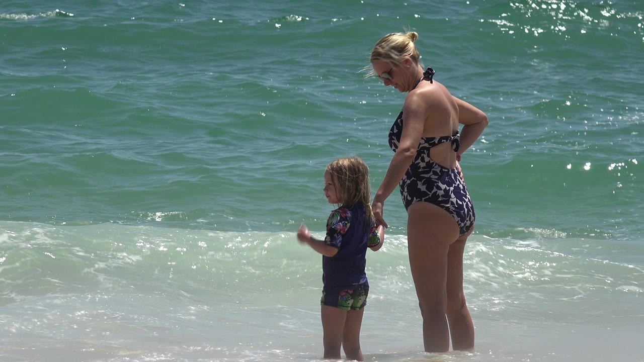 Mother and young daughter stand in shallow ocean surf.