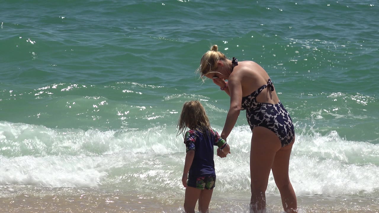 Woman holds a child's hand at the edge of ocean surf.