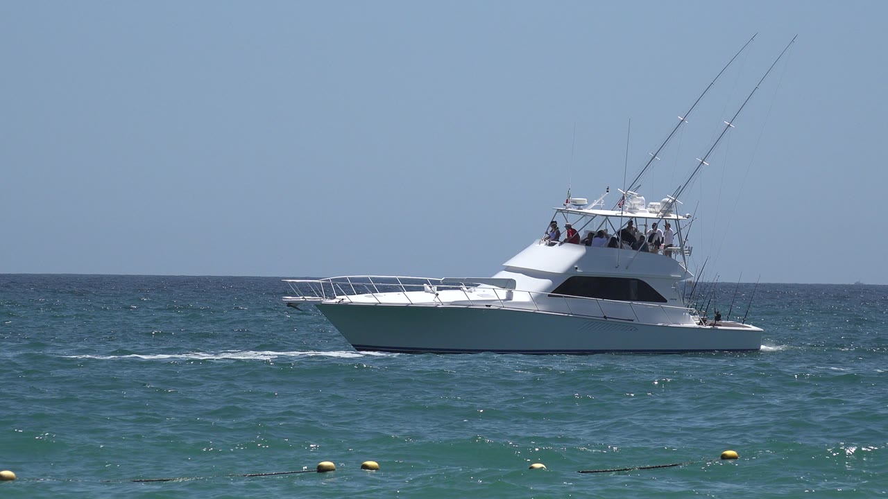 The yacht remains broadside on the Cabo San Lucas water, with dark cabin windows, several indistinct people on the flybridge, and small wake lines behind the hull.