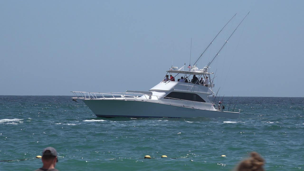 A white sportfishing boat sits offshore in Cabo San Lucas, Mexico, with fishing rods above the cabin, small waves across the water, and partial beachgoers cropped along the bottom edge.