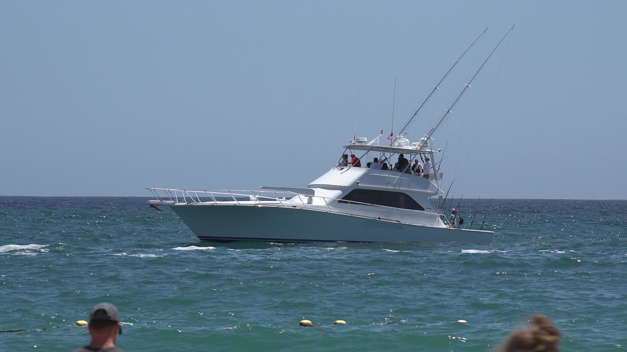 The boat remains centered offshore in Cabo San Lucas, Mexico, with darker blue water behind it, small white wave breaks in front, and cropped heads at the lower frame edge.