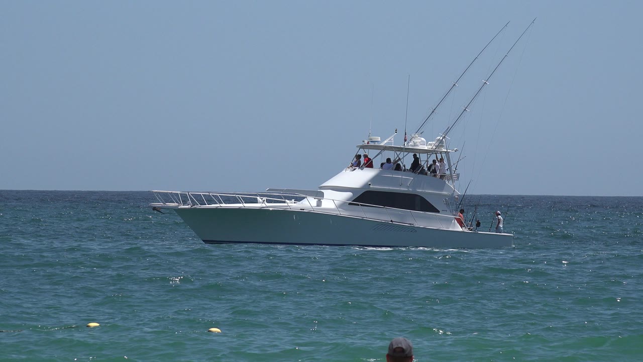 White yacht in Cabo San Lucas, Mexico, sits near center frame on choppy blue-green water, with small figures on the deck and tall fishing rods silhouetted against clear sky.