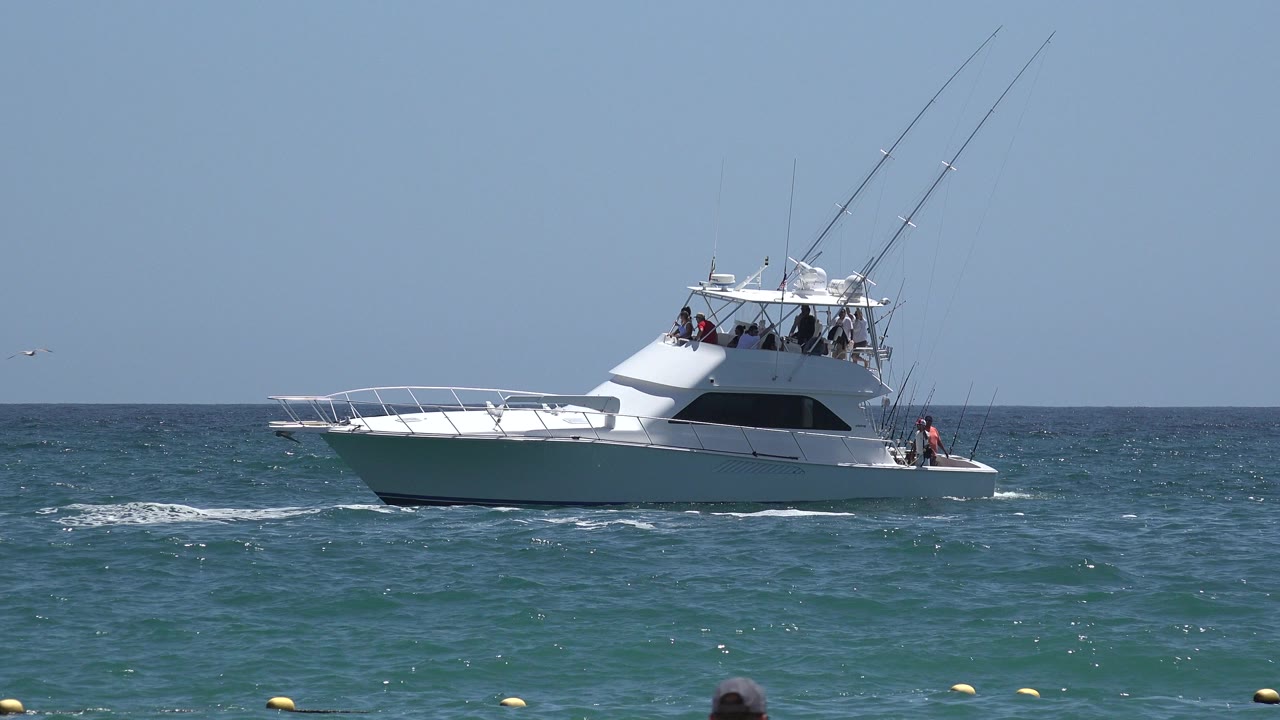 White yacht on blue-green ocean with people aboard and buoys near the foreground.