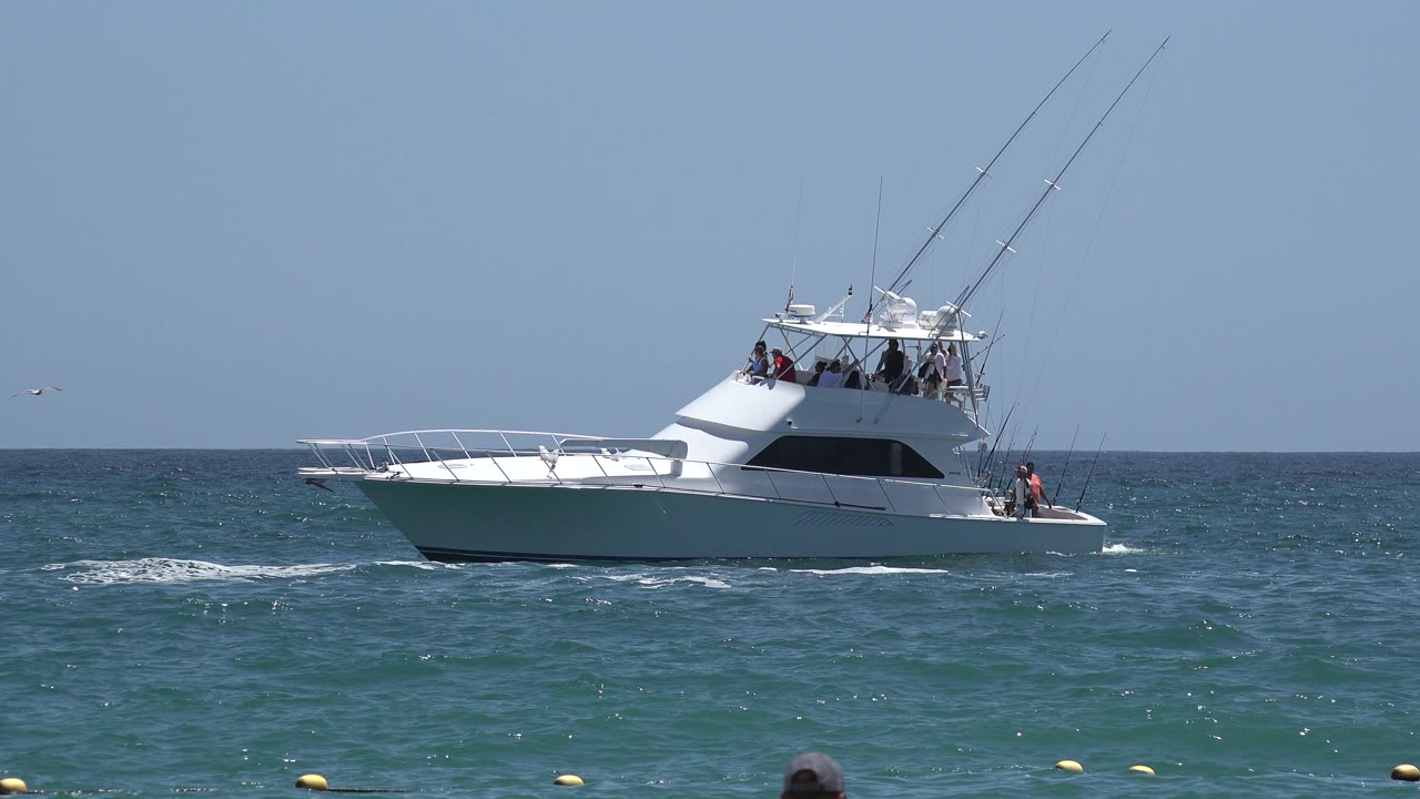 White sport fishing yacht in Cabo San Lucas, Mexico, remains in profile as small waves break along the hull and people stand on the flybridge and rear deck.