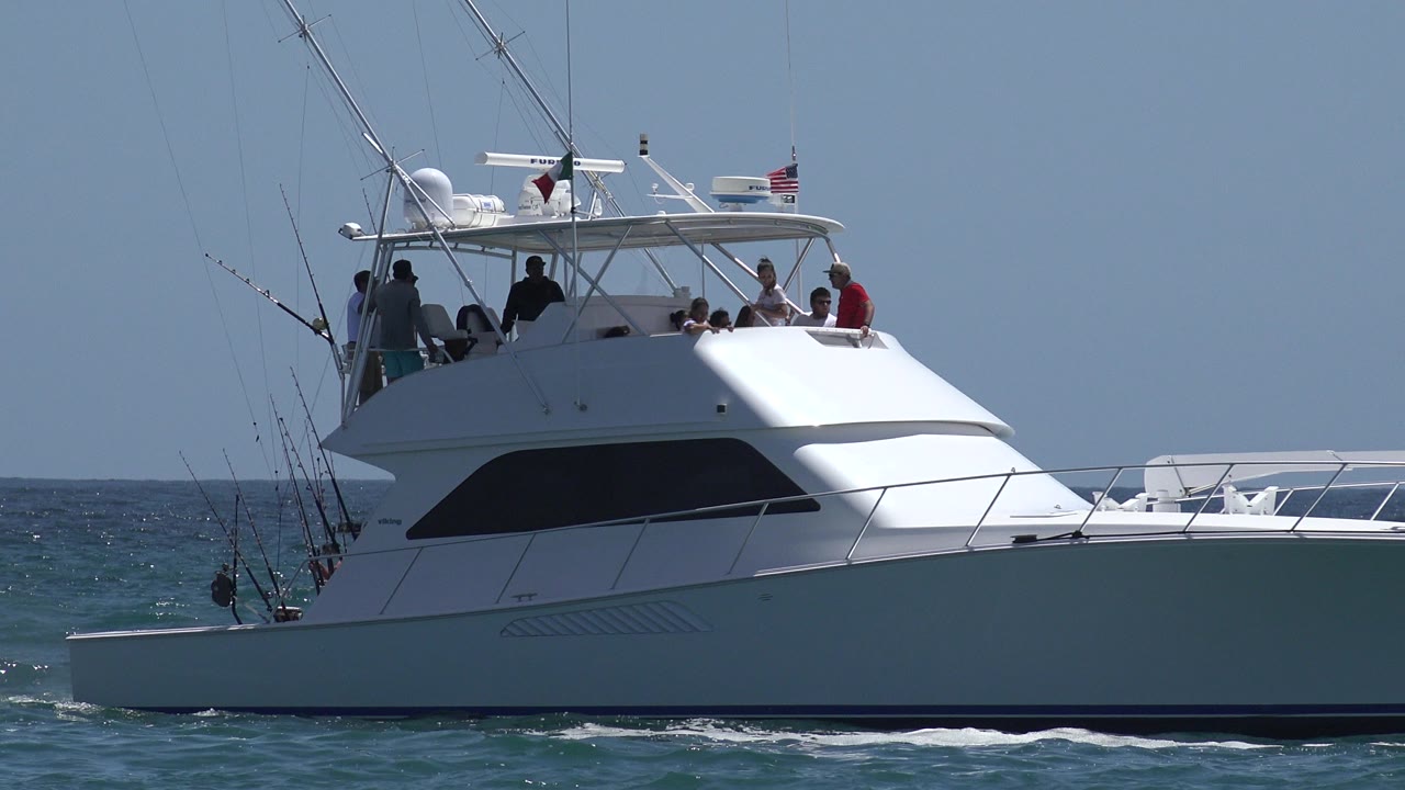 A white sport fishing boat in Cabo San Lucas, Mexico, moves across blue water with several people visible on the upper deck and fishing rods angled from the stern.