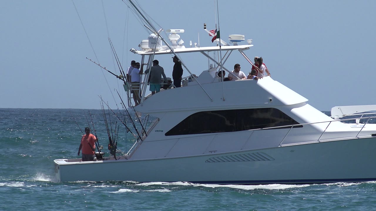 A white sportfishing boat in Cabo San Lucas, Mexico, crosses blue open water with several adults on the upper deck and a man standing near the stern.