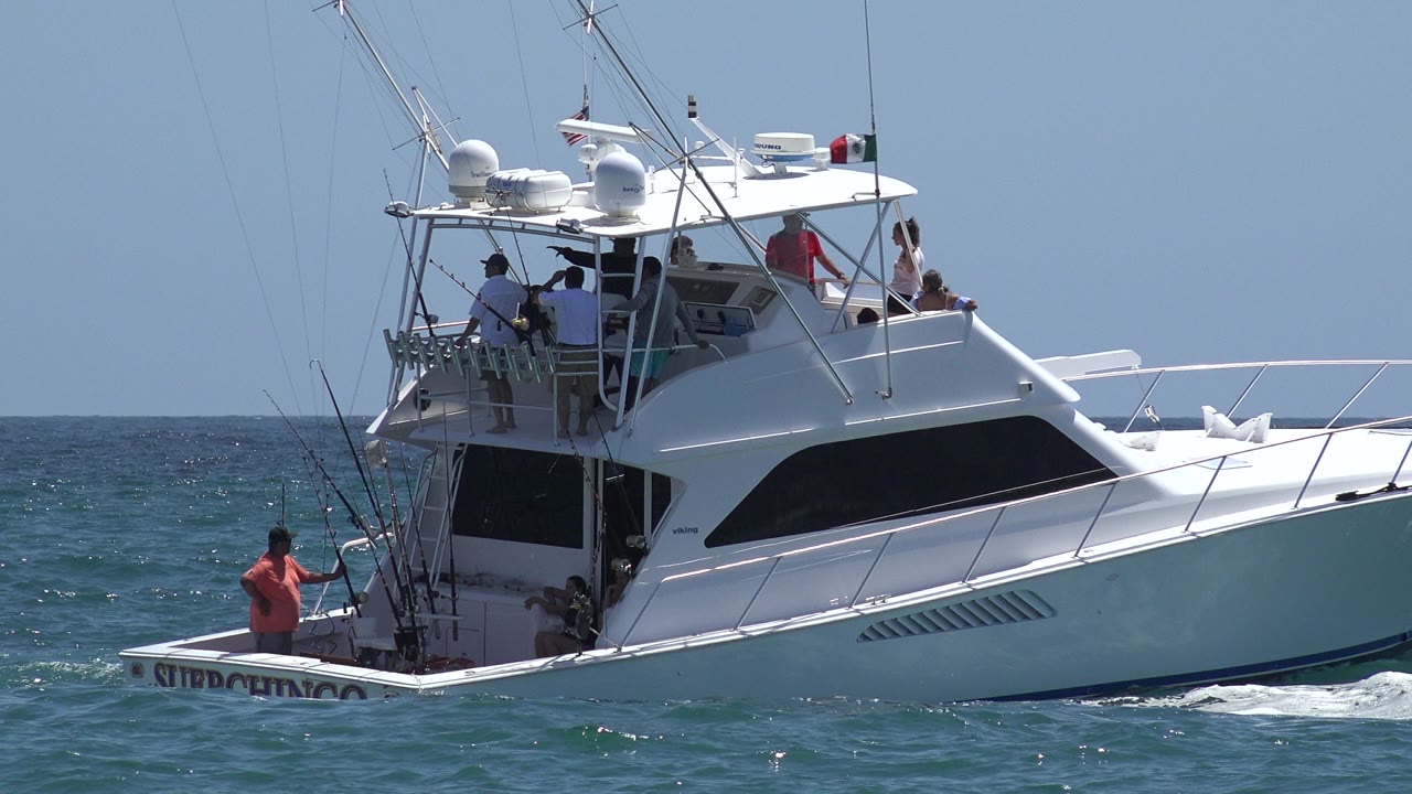 The Cabo San Lucas sportfishing boat remains broadside on the water, with several people gathered on the flybridge and a person in a red shirt standing near the stern rods.