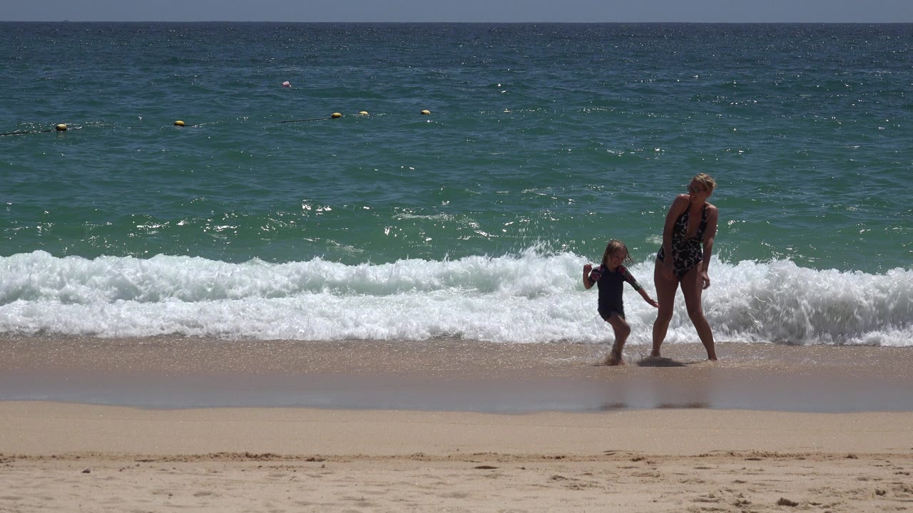 In Cabo San Lucas, Mexico, a woman and child hold hands on wet sand while a low white wave breaks behind them under bright sun.