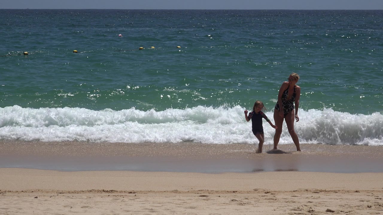 In Cabo San Lucas, Mexico, the woman and child walk along the shoreline as foamy surf spreads across the wet sand behind their feet.