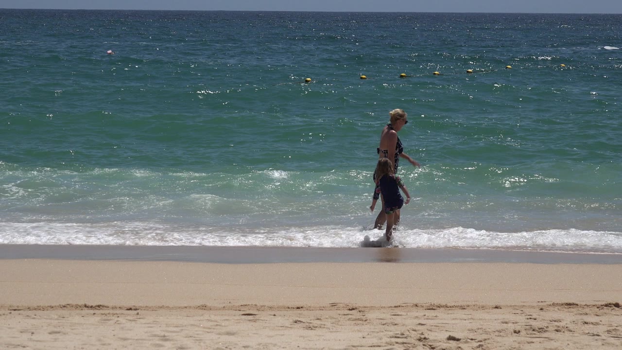 Woman and child walking through shallow surf on a sandy beach.