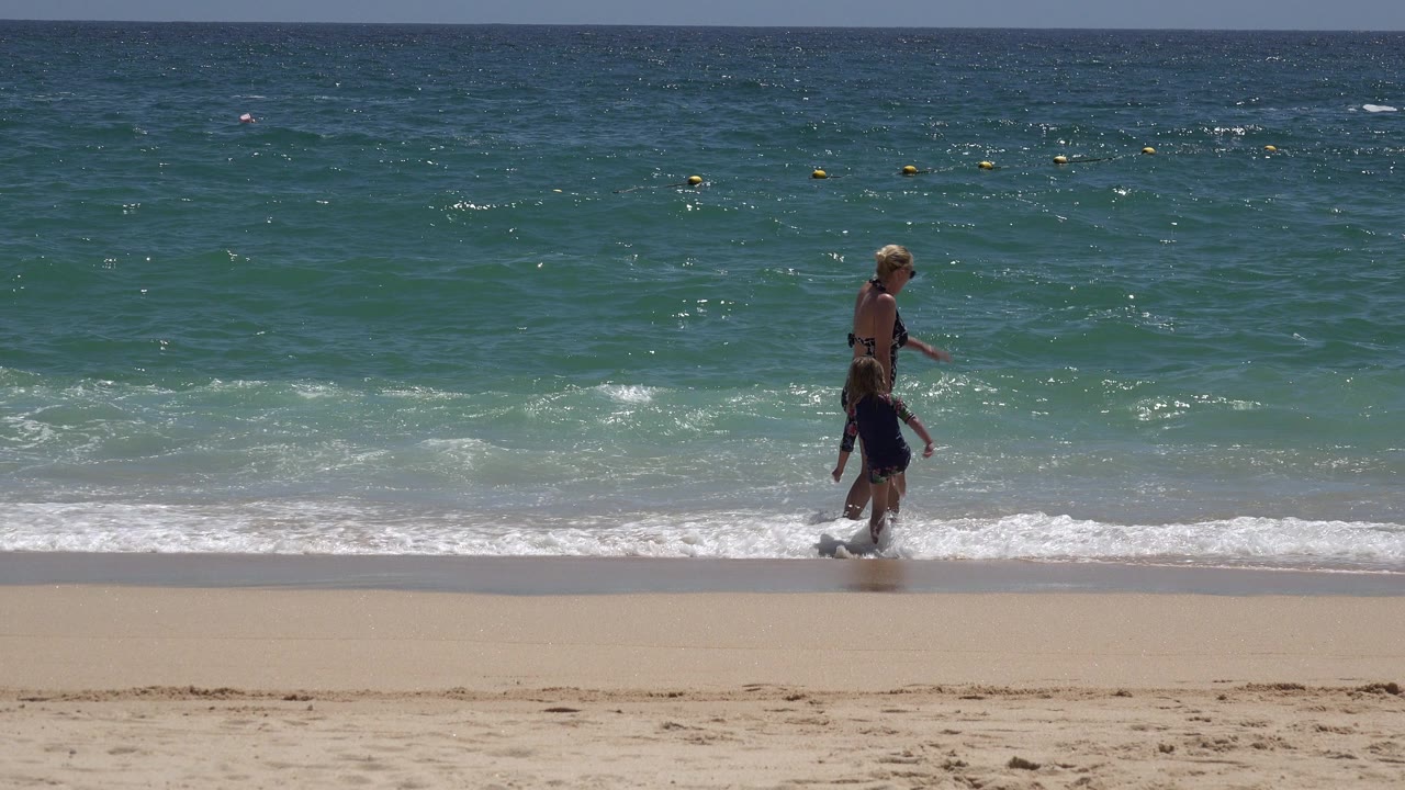 The adult and child remain near the waterline in Cabo San Lucas as low white surf breaks behind them and the ocean fills the background.