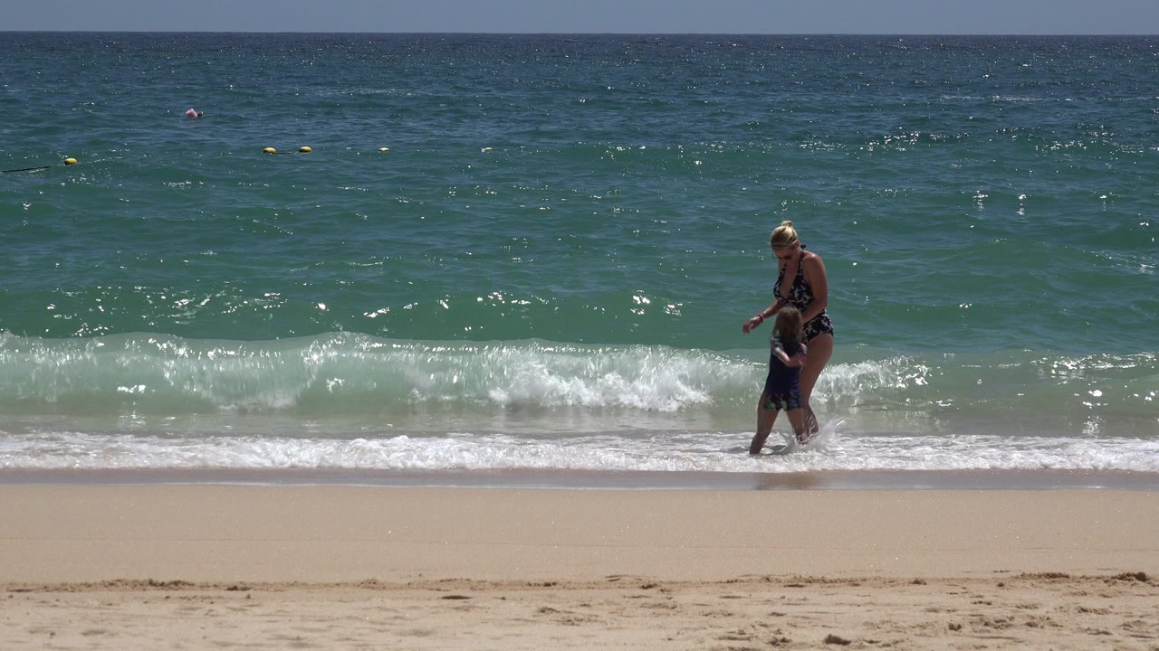 Adult woman and child standing at the edge of breaking surf.